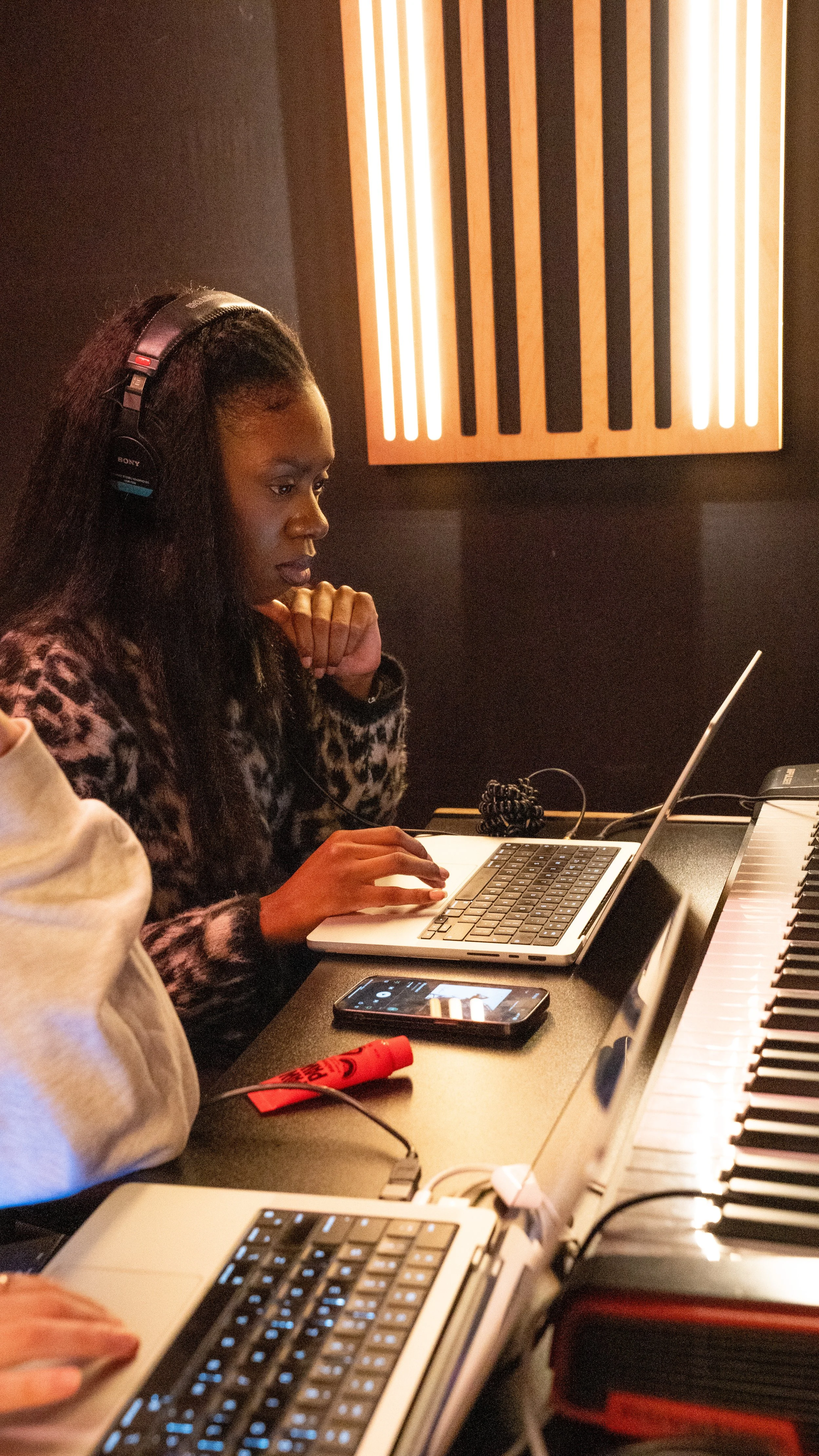 A woman wearing headphones working on a laptop in a recording studio, with a keyboard and a smartphone on the desk.