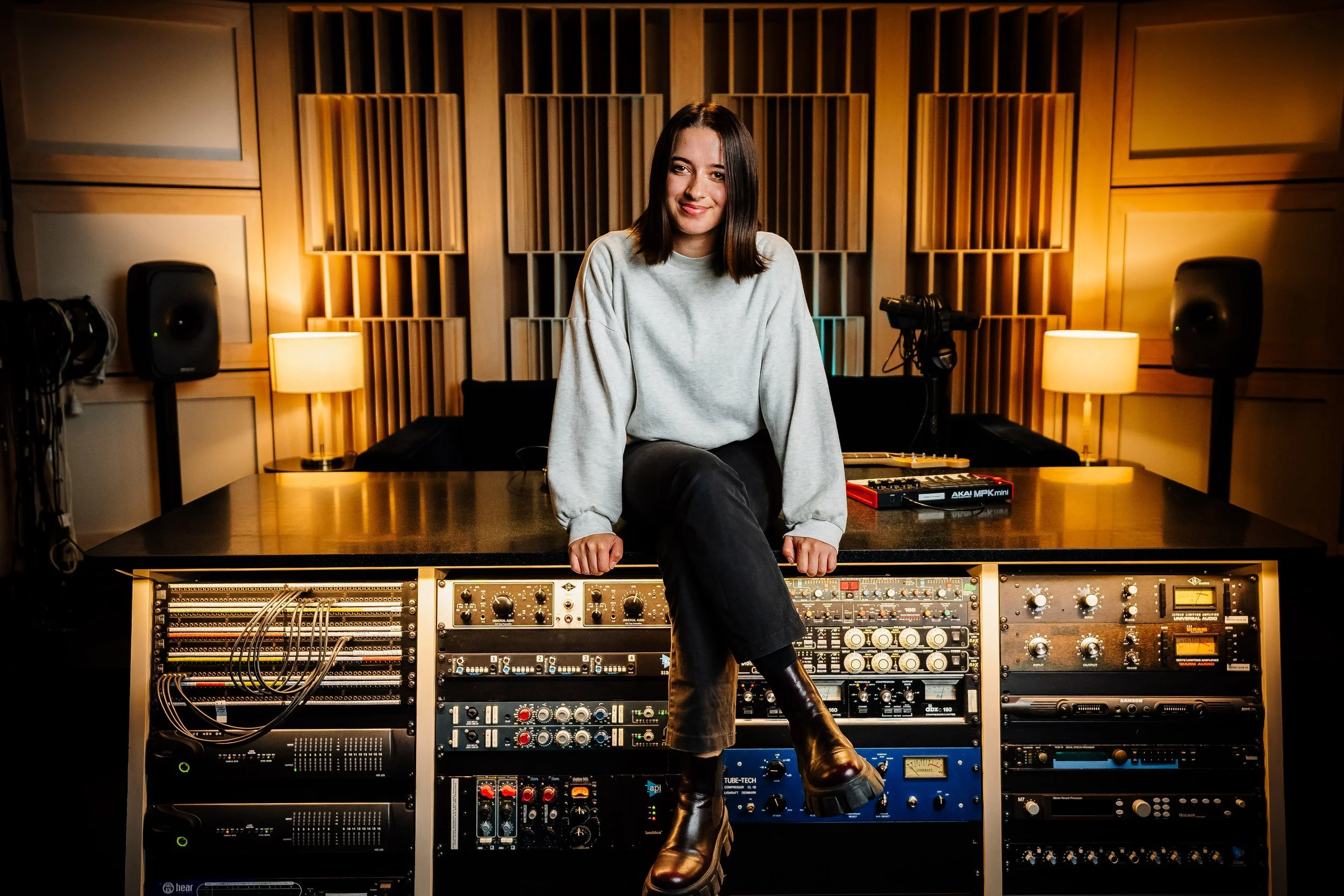 A young woman in a gray sweatshirt and dark pants sitting on audio equipment in a recording studio, smiling at the camera with her leg crossed, surrounded by soundproof panels and lighting.