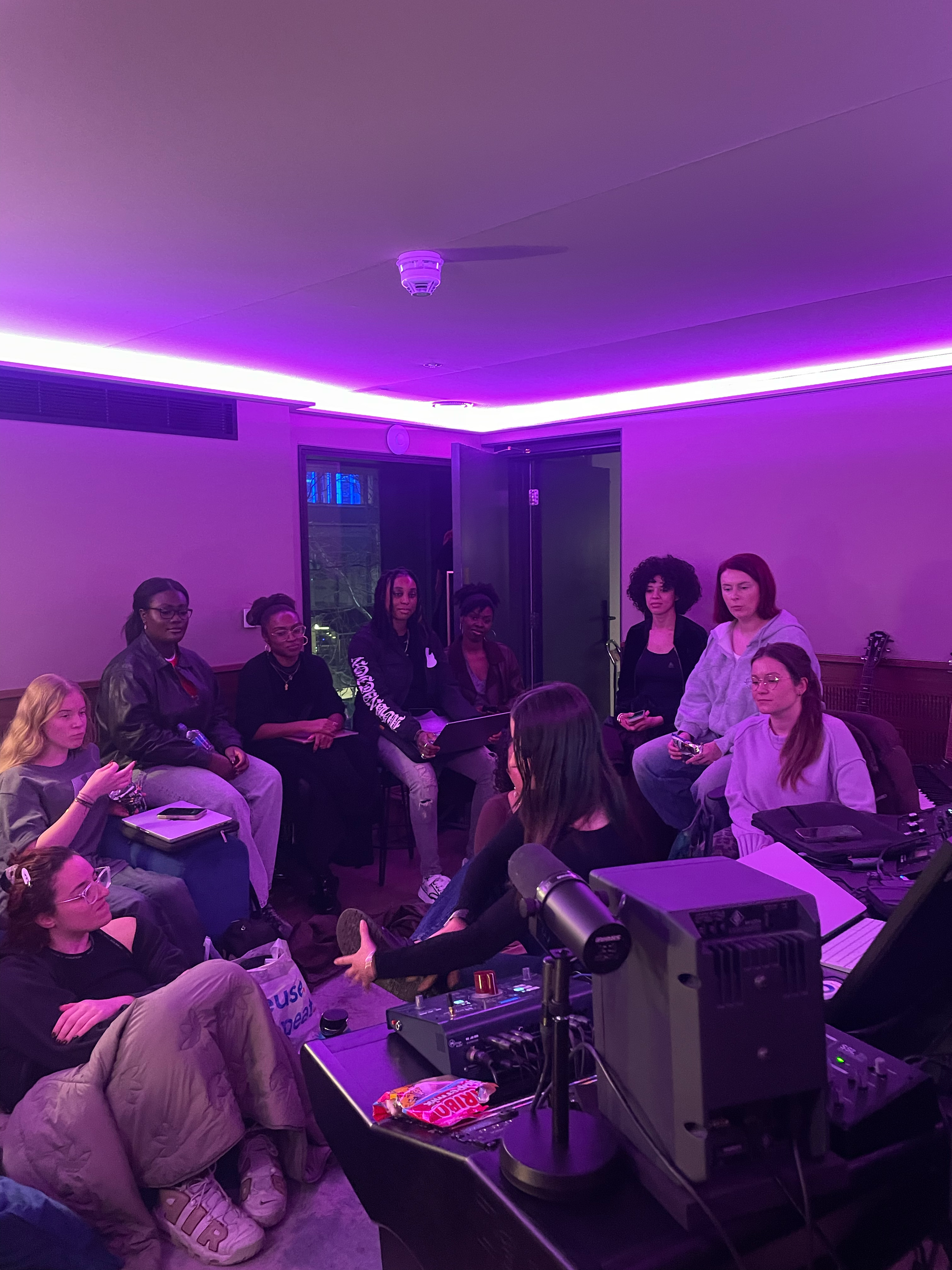 Group of women sitting in a circle in a room illuminated with purple neon lighting, with a DJ setup in the foreground.