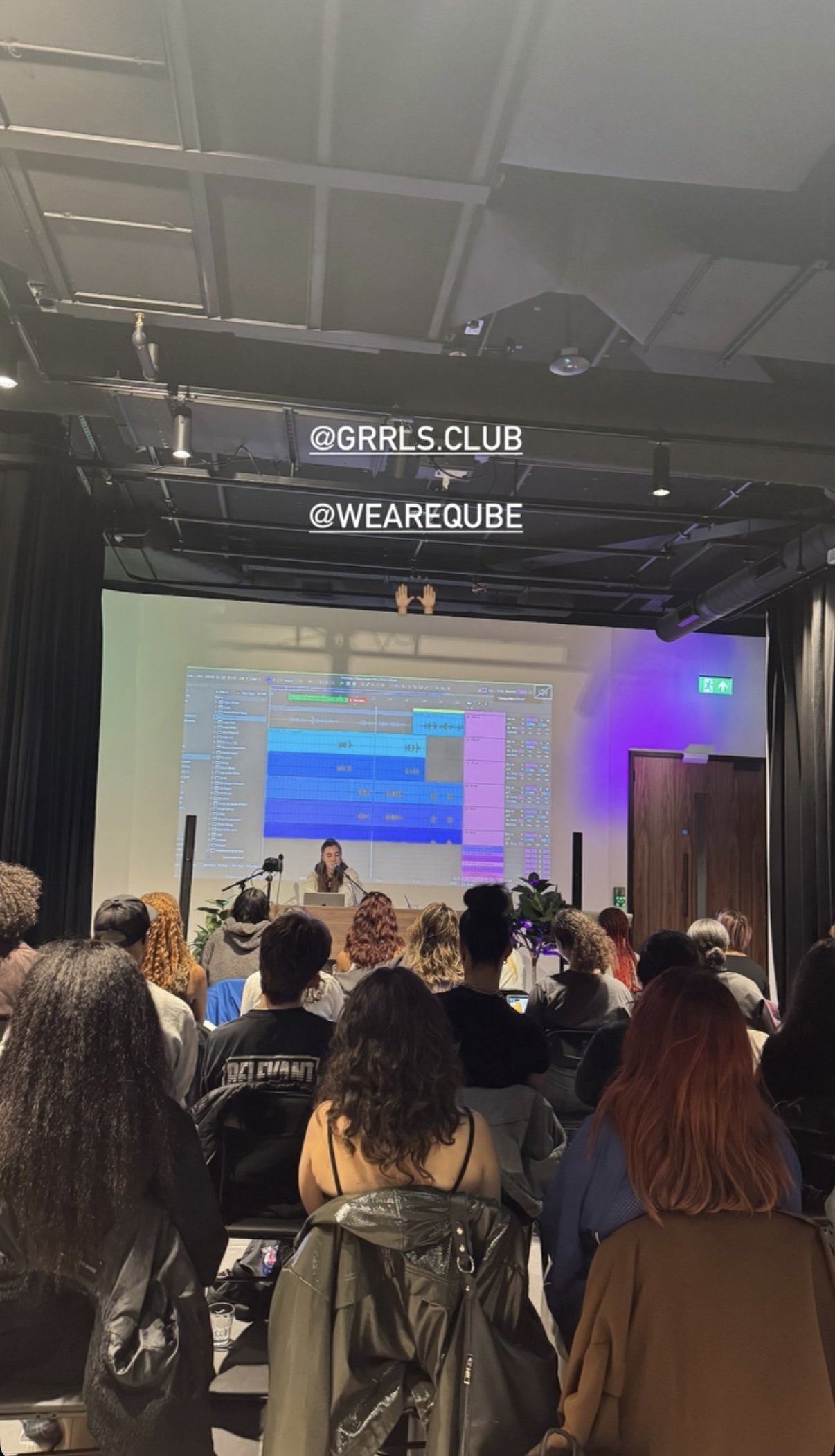 A woman presenting on stage at a music production workshop or seminar, with digital audio workstation software displayed on a large screen behind her, and an audience of people seated and listening.
