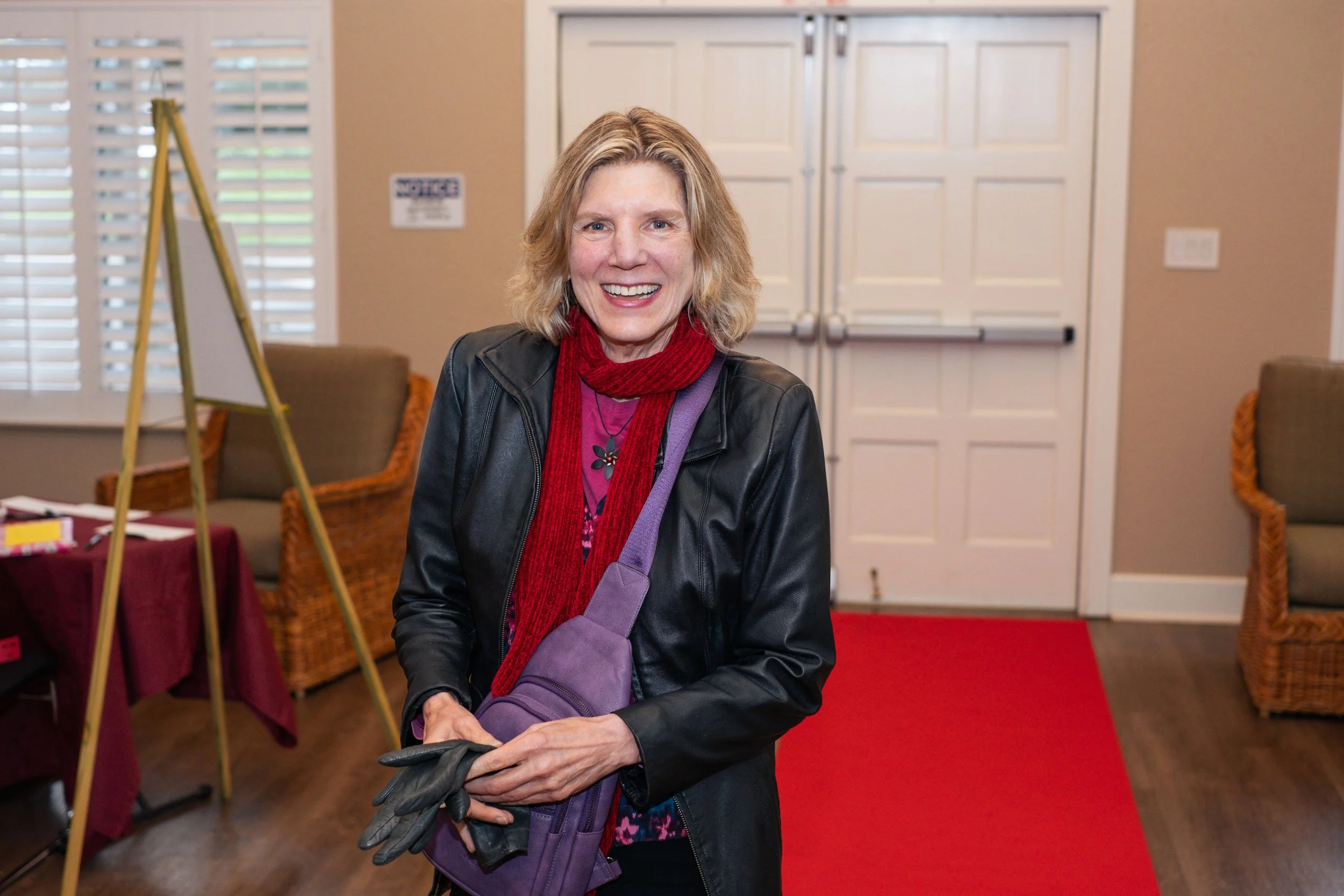 A woman with shoulder-length blonde hair smiling, wearing a black leather jacket, burgundy scarf, purple shirt with floral design, carrying purple shoulder bag, holding gardening gloves in her hands, standing in a room with a red carpet, wicker chairs, and a closed white door in the background.