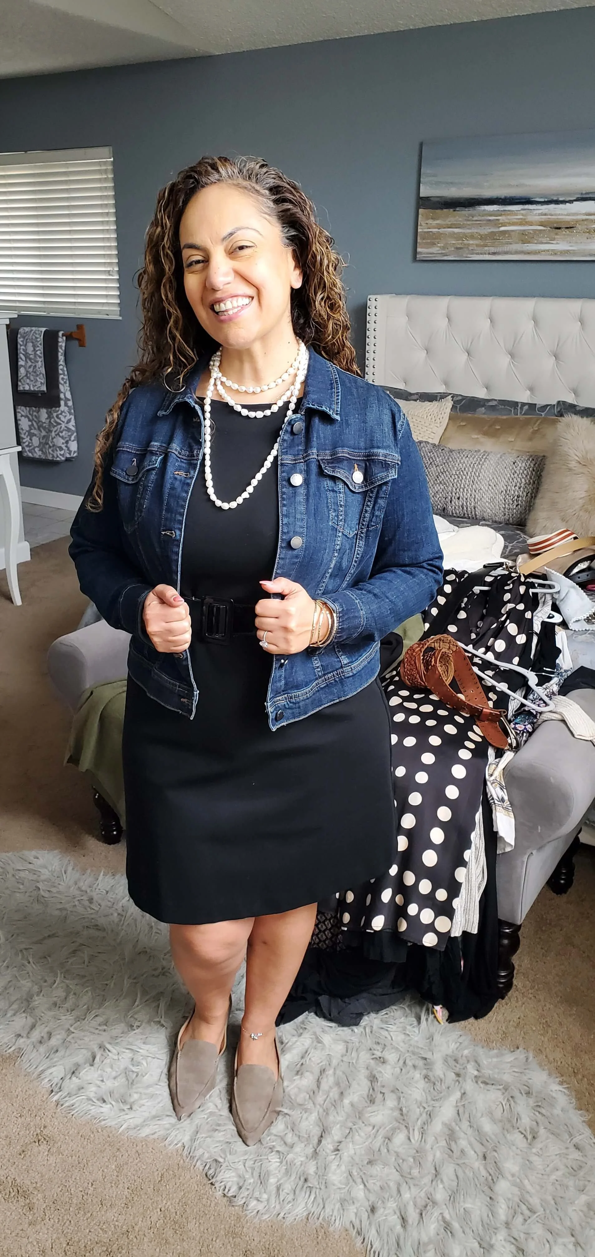 A woman standing in a living room, smiling at the camera, wearing a black dress with a black belt, a denim jacket, beige flats, and multiple pearl necklaces. There is a bed with clothing and accessories behind her, a window with blinds, and a painting on the blue wall.
