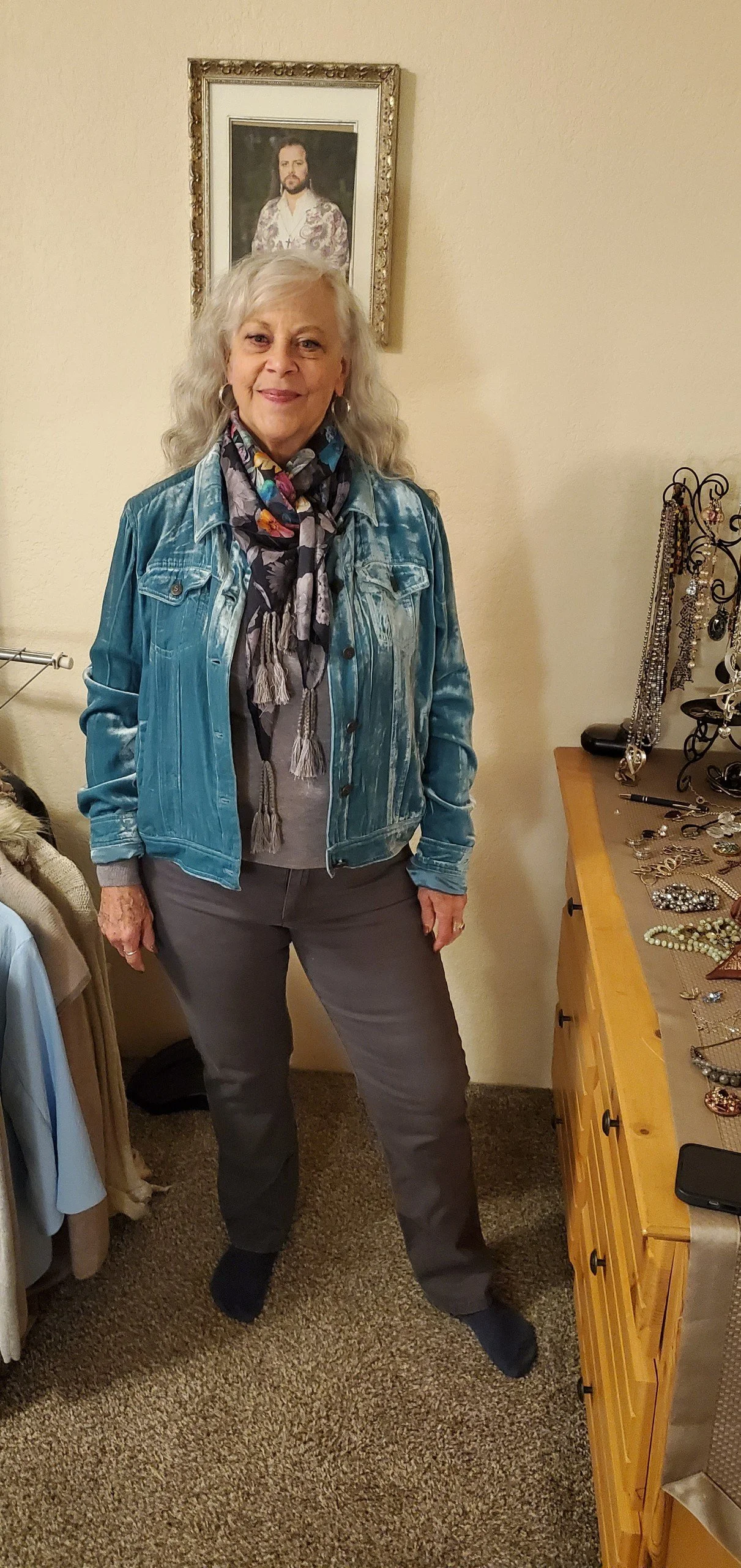 An older woman with gray hair standing indoors, wearing a bright blue denim jacket, gray pants, and a colorful scarf. Behind her on the wall is a framed portrait of a man with long hair and a beard.