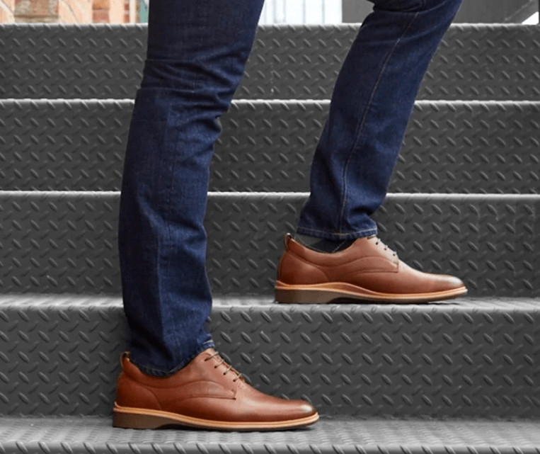 A person's legs and feet wearing dark blue jeans and brown leather shoes, standing on gray industrial stairs.
