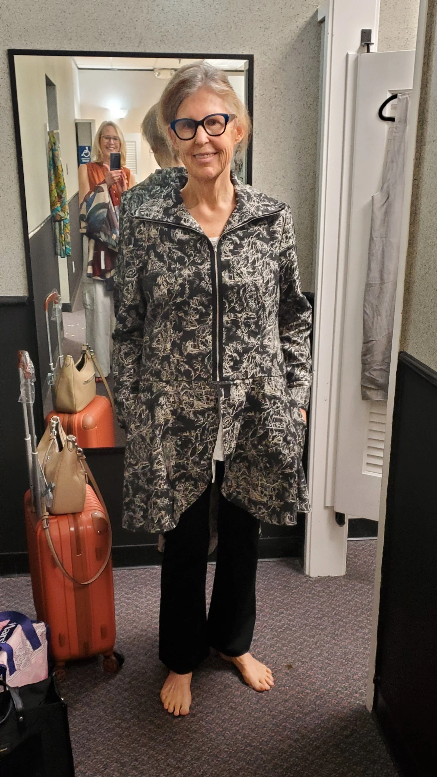 A woman with glasses and gray hair taking a photo in a mirror, wearing a black and gray patterned coat and black pants, standing barefoot in a hotel room.