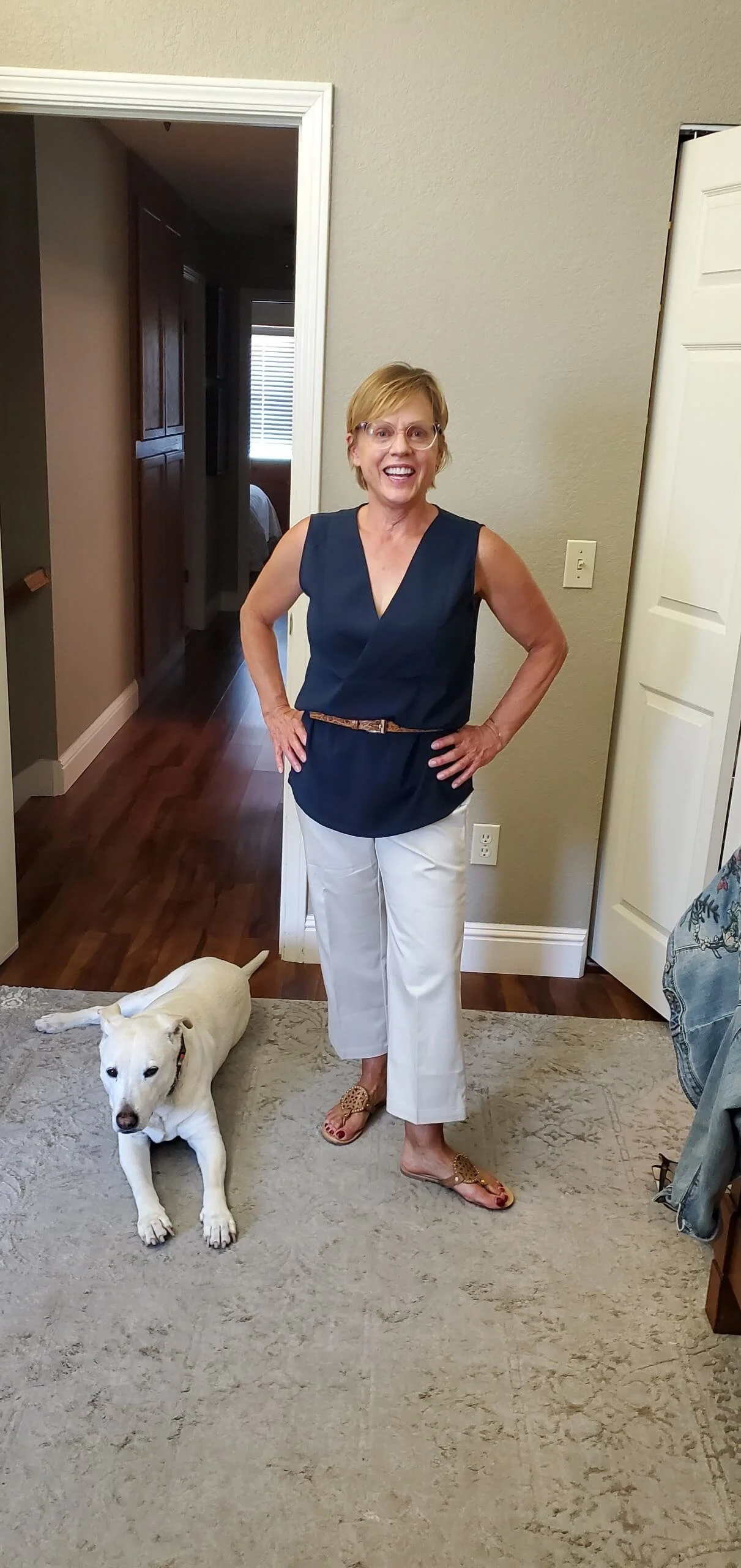 A woman with short blonde hair, wearing glasses, a sleeveless navy blue top, white pants, and sandals, standing indoors with hands on hips and smiling. A white dog is lying on the carpeted floor beside her.