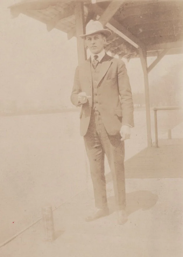 Significantly faded circa 1910 image of man in hat and suit standing on pier.