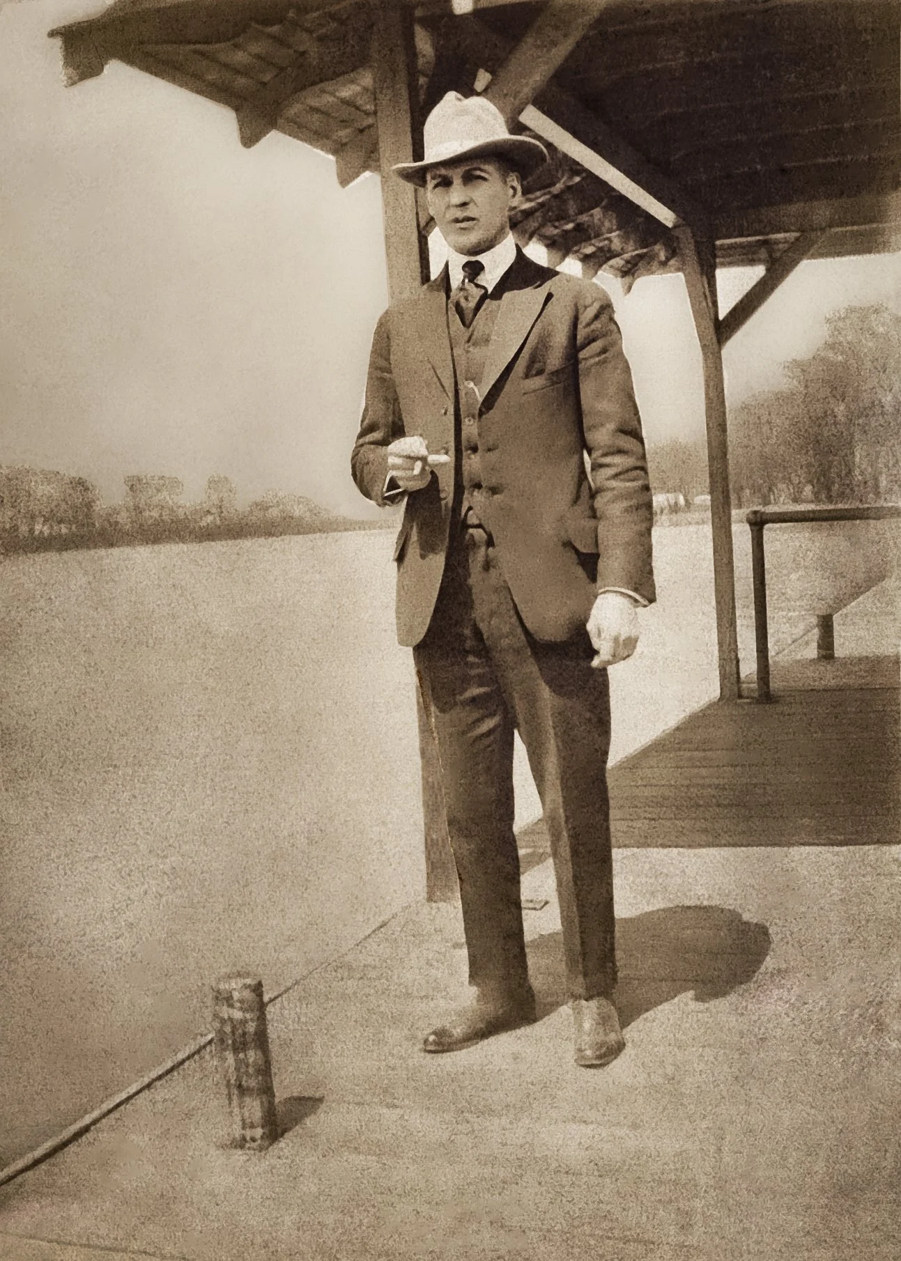 Crisp c. 1910 image of man in hat and suit standing on pier, with trees in the far background.