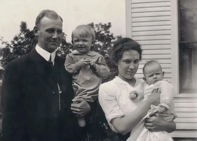 Black and white photo of a man, a woman, and two young children outside a house, with trees in the background.