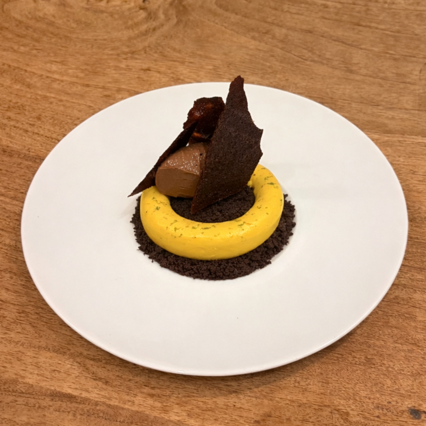 A plated dessert with a yellow circular base, a dark chocolate crumb, and garnishes of chocolate elements and dark crispy tuile on a white plate.