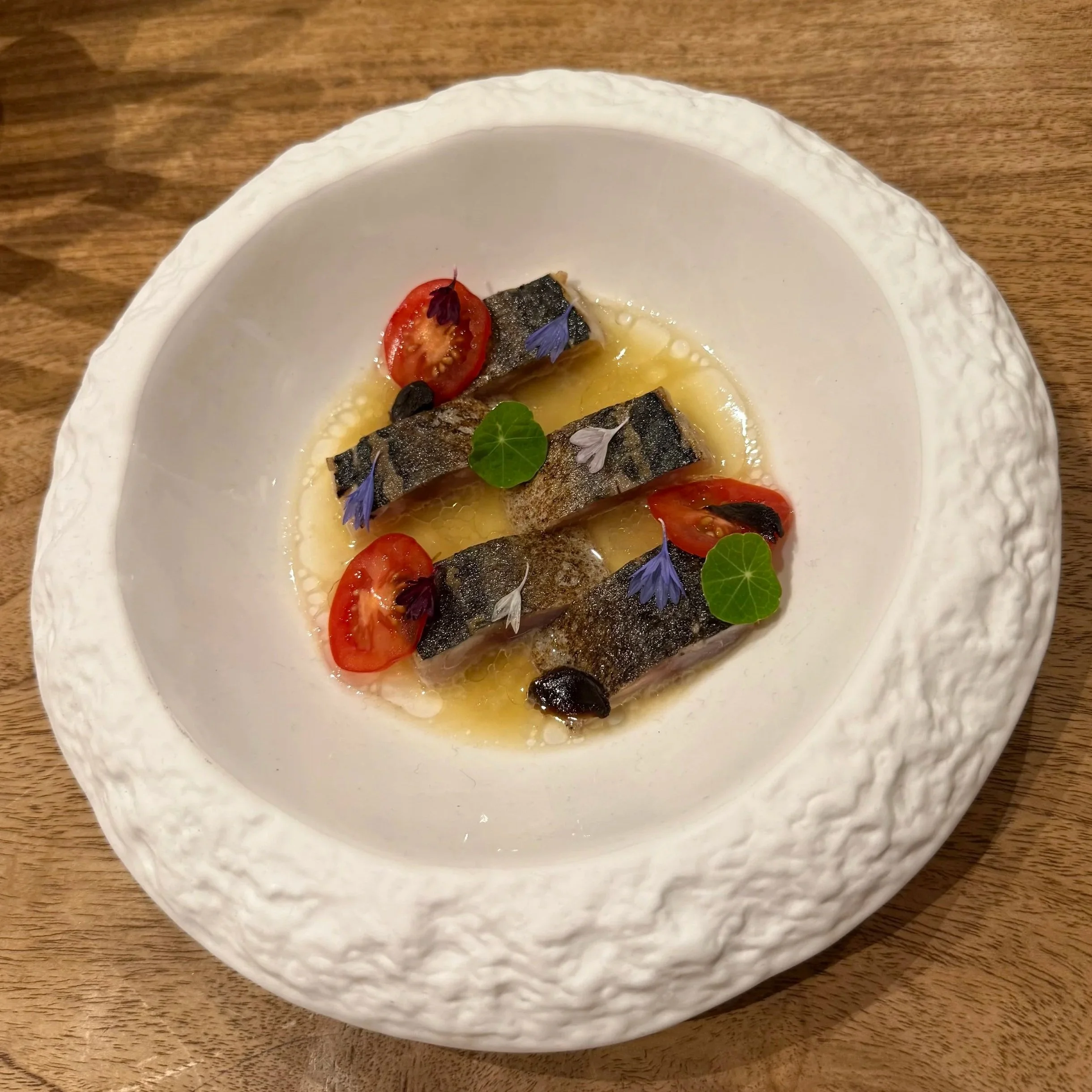 A white decorative bowl with textured edges on a wooden surface, containing a dish garnished with cherry tomatoes, edible flowers, and herbs.