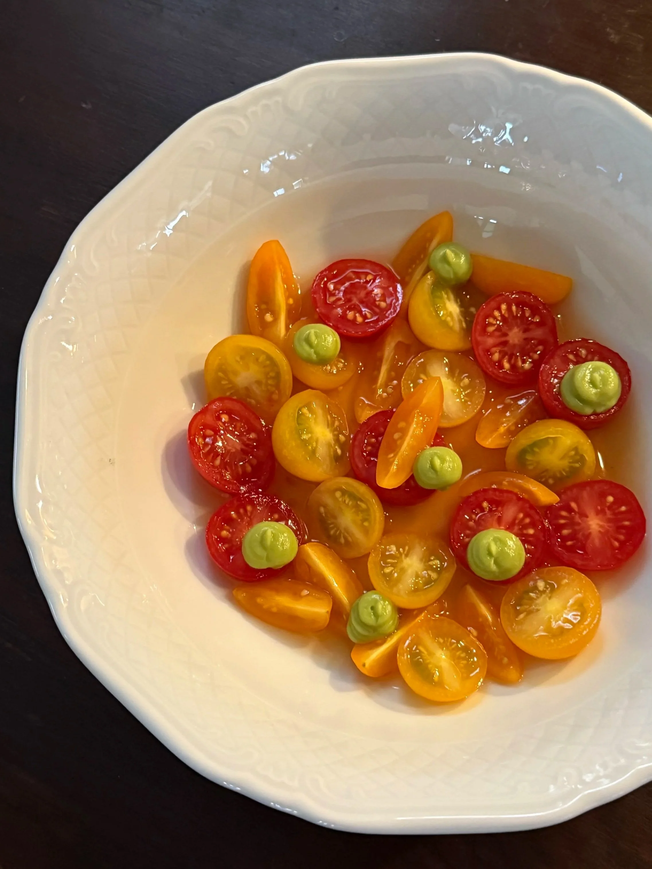 A white bowl with a textured rim holding a colorful assortment of halved cherry tomatoes and small green peas, placed on a dark wooden surface.