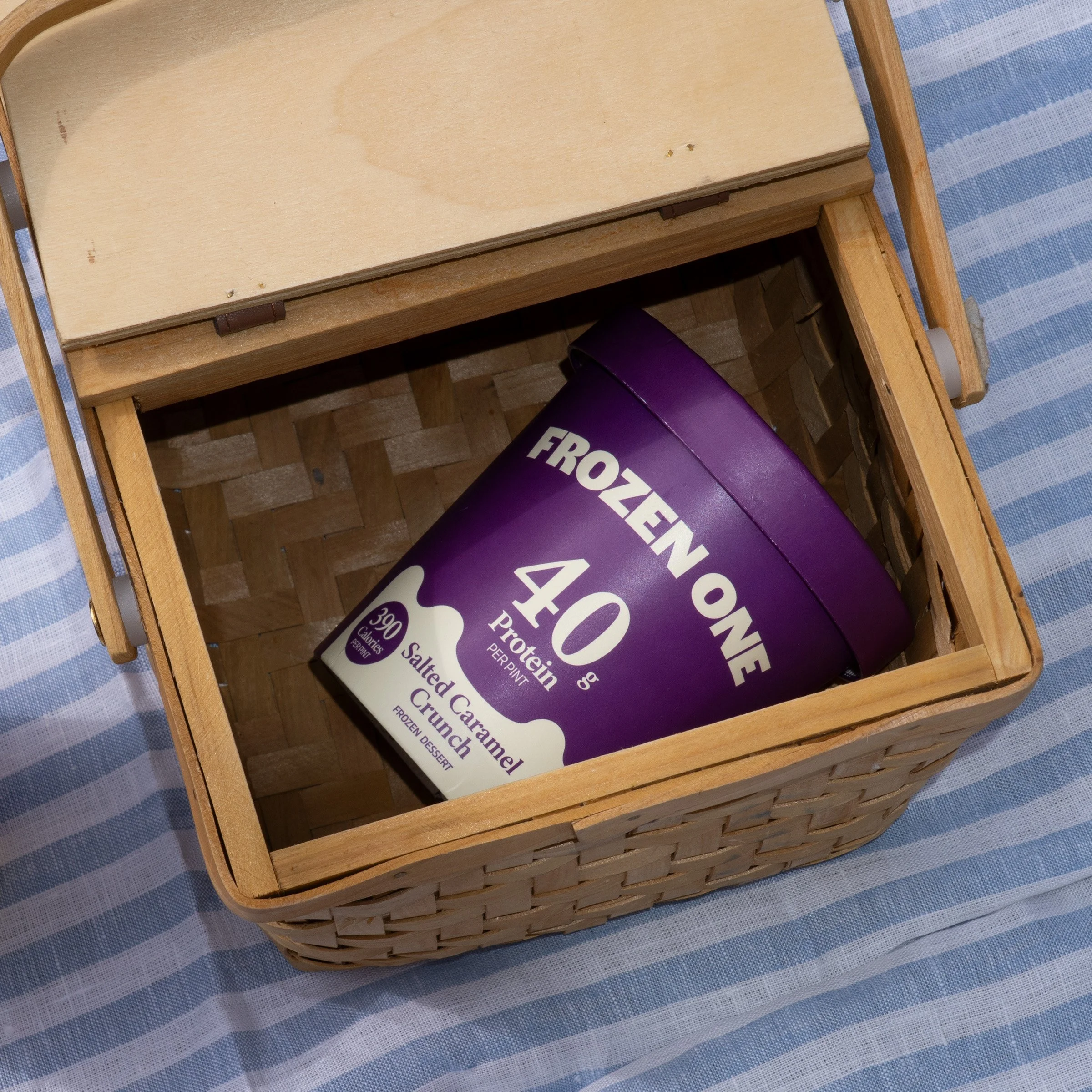 A wooden basket containing a purple container of frozen crumble dessert on a blue and white striped fabric surface.