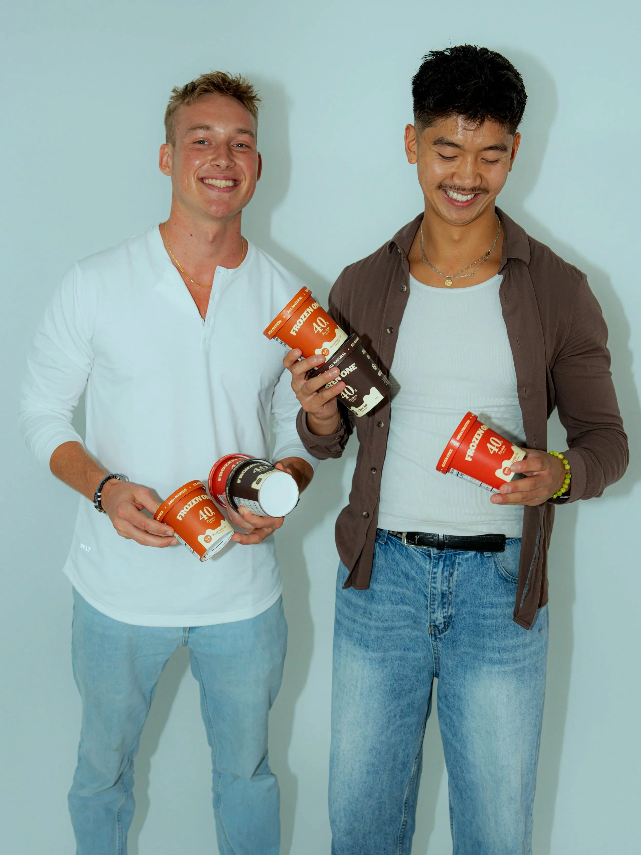 Two young men standing against a plain wall, each holding containers of frozen yogurt, smiling and looking down.