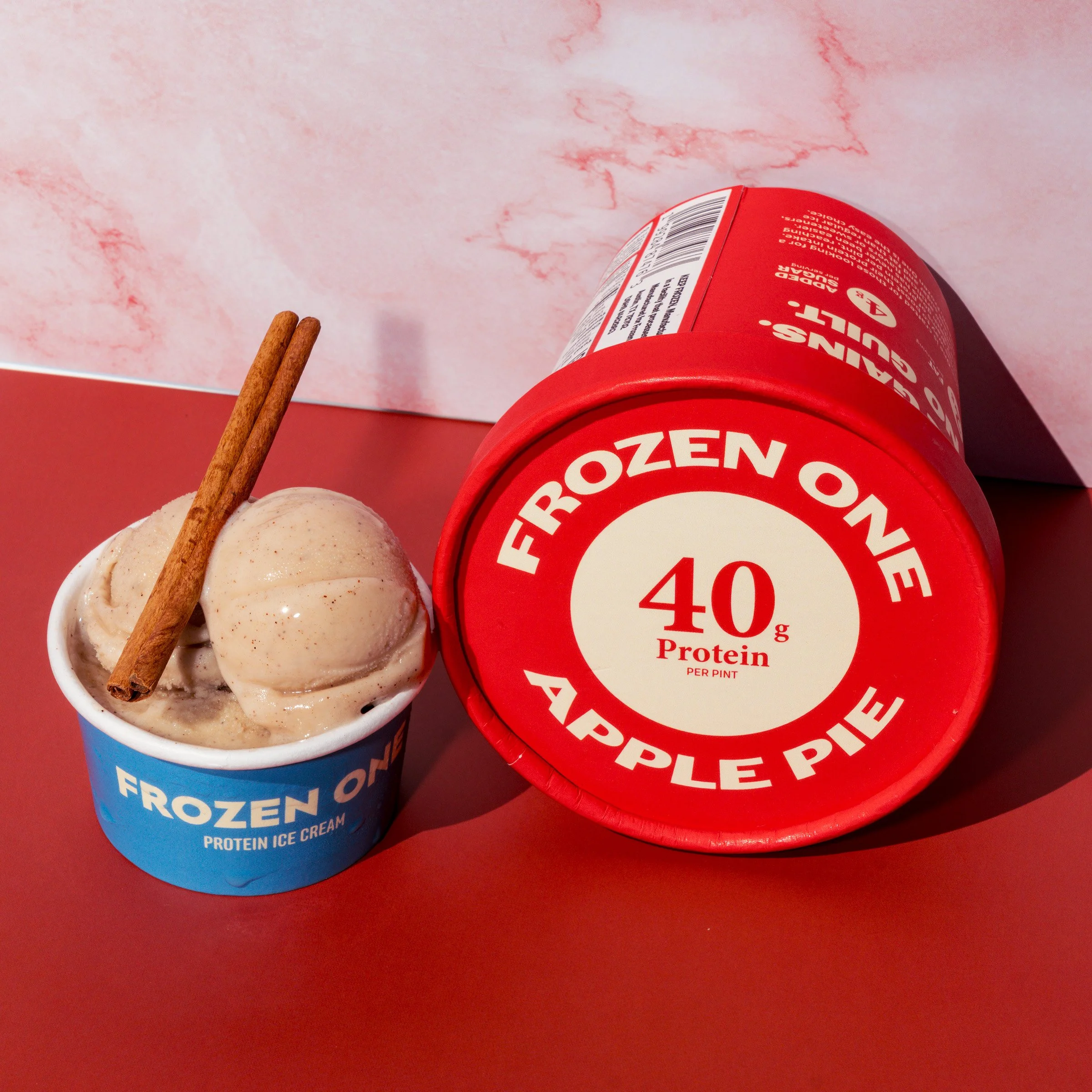 Two containers of frozen protein ice cream, one blue and one red, with cinnamon sticks on top, placed on a reddish surface against a pink marbled background.