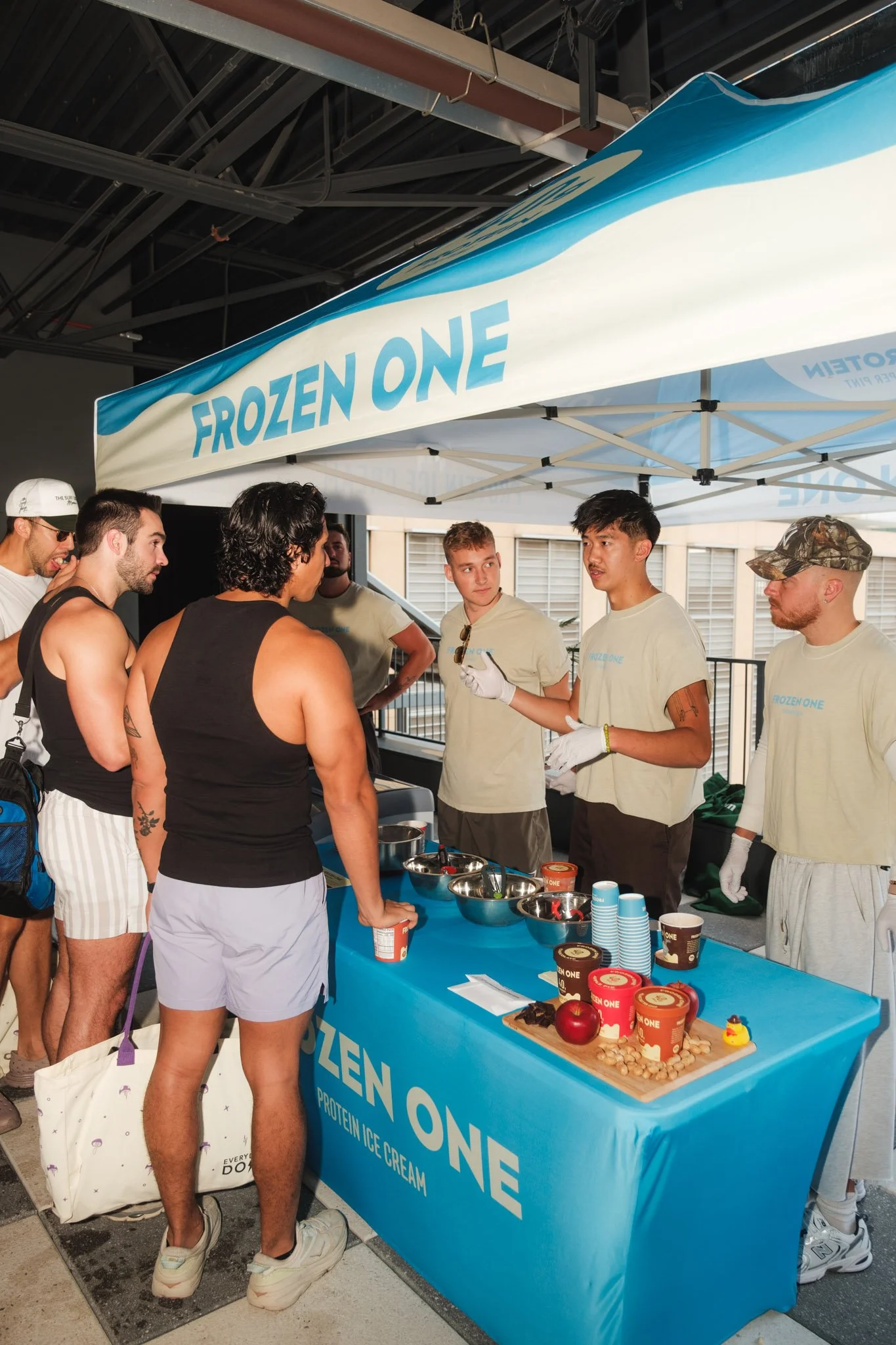 Group of people at a Frozen One ice cream booth, with staff in branded t-shirts interacting with attendees, under a canopy with promotional items on a table.