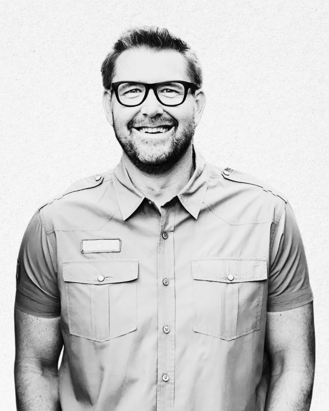 Black and white portrait of a smiling man wearing glasses and a collared shirt with pockets, standing against a plain background.