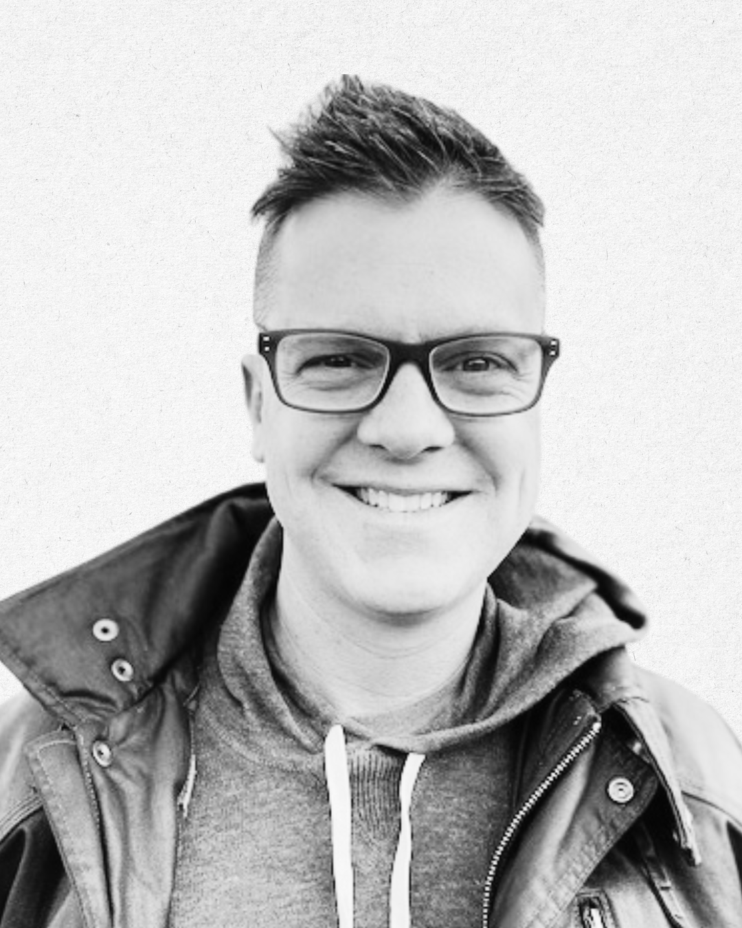 Black and white photo of a smiling man wearing glasses, a hoodie, and a jacket, with short styled hair.