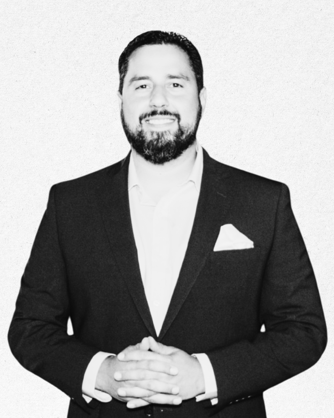 Black and white portrait of a man with dark hair and beard, wearing a suit jacket, white shirt, and pocket square, smiling with hands clasped in front of him.