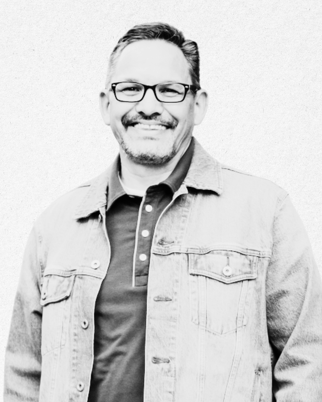 A smiling man with glasses and a goatee, wearing a denim jacket over a polo shirt, standing against a plain background.