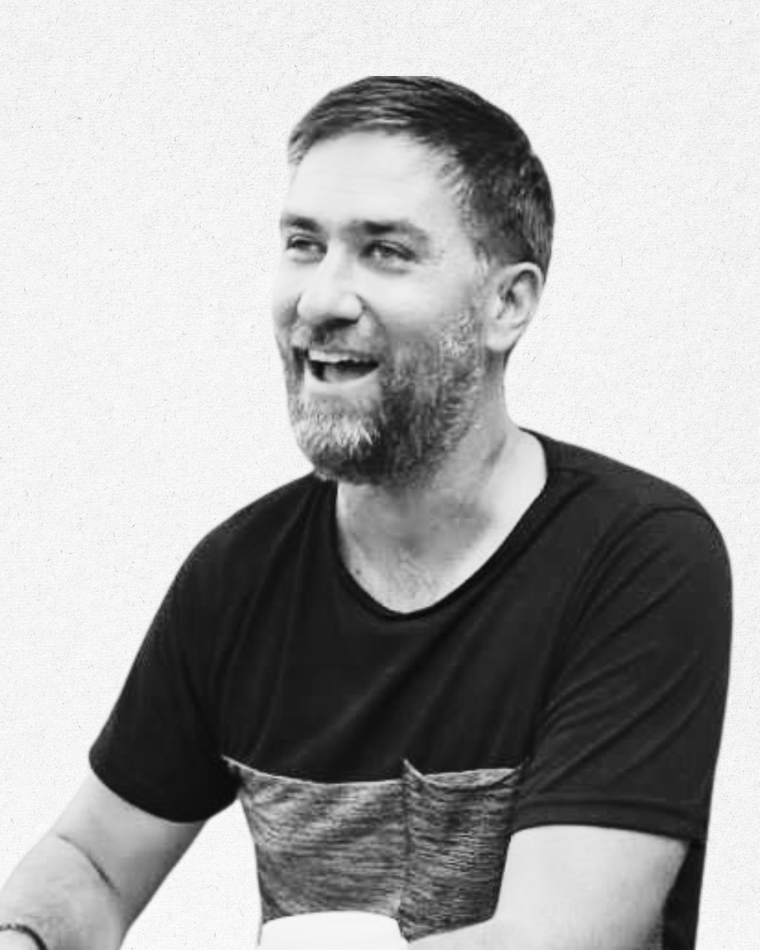 Black and white photo of a smiling man with a beard, wearing a dark t-shirt with a textured pocket detail, against a plain background.
