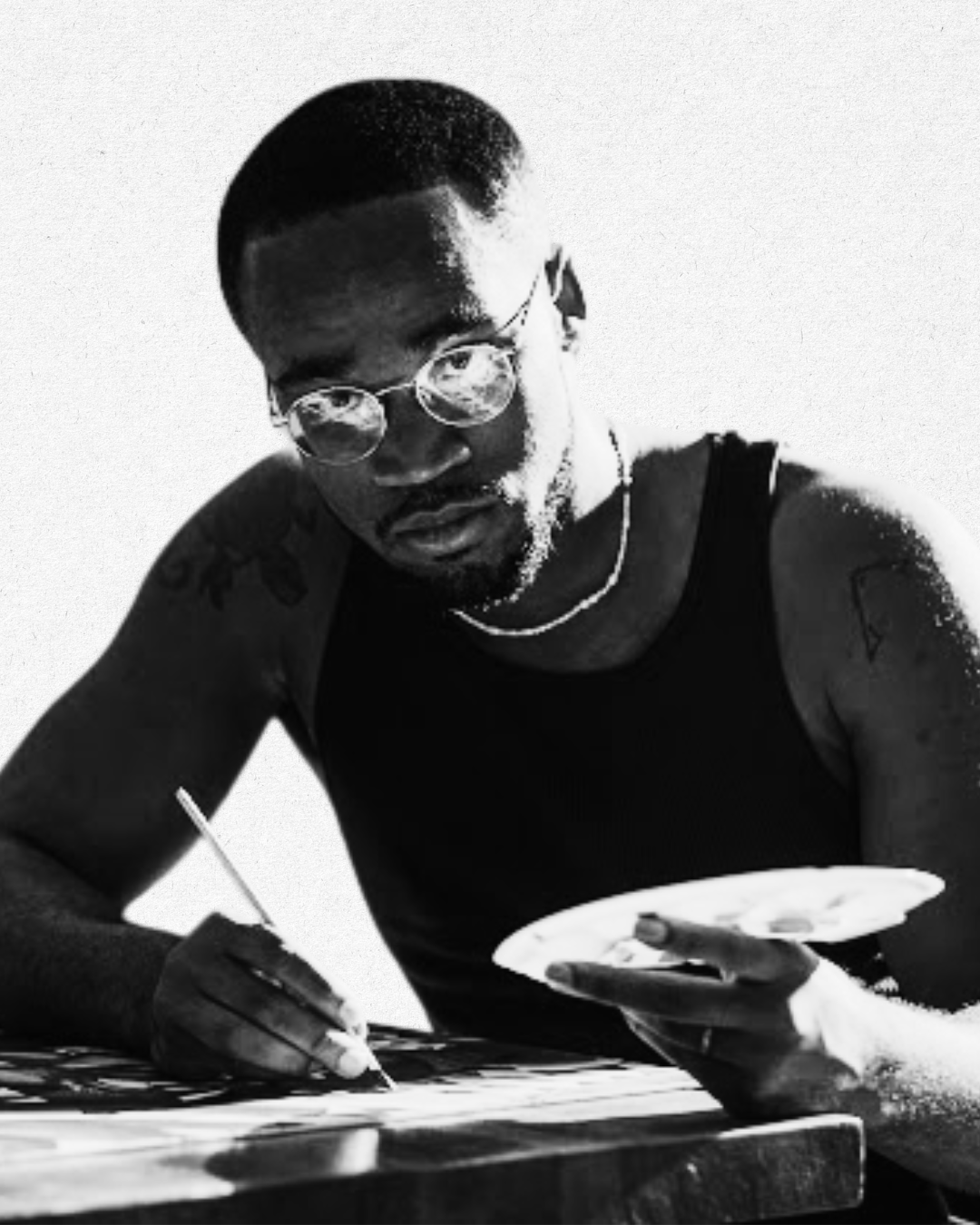 A young man with glasses and tattoos, wearing a sleeveless shirt and necklace, is painting on a canvas with a brush and holding a palette in his other hand. Black and white photo.