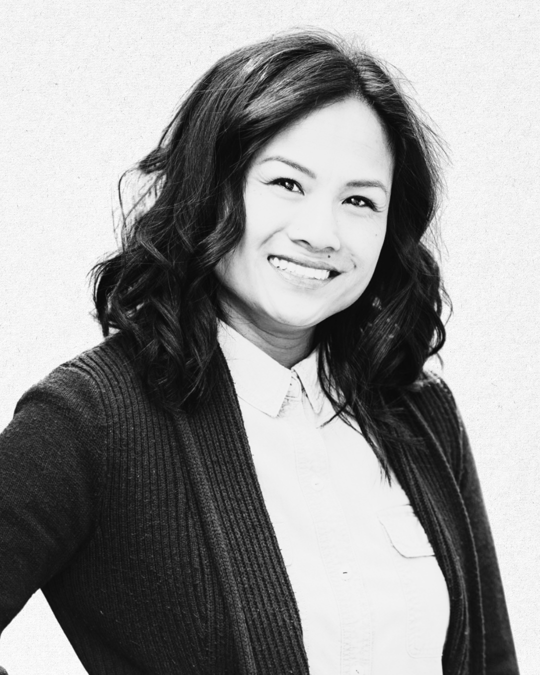 Black and white portrait of a woman with wavy hair smiling, wearing a white collared shirt and a dark cardigan.