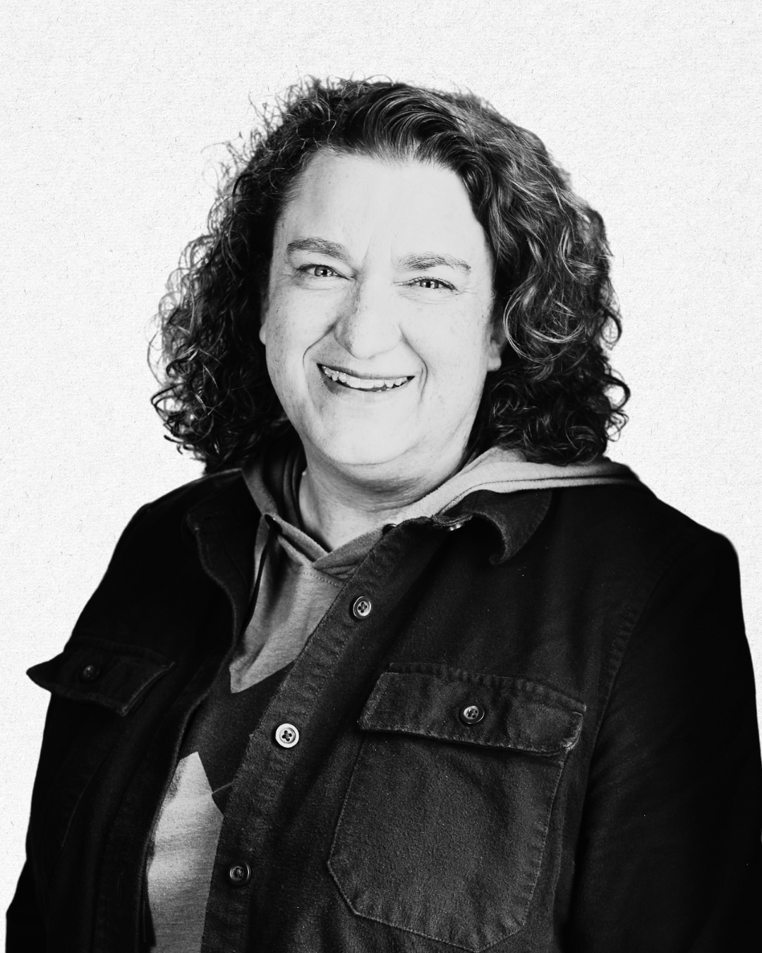 Black and white portrait of a person with curly hair smiling at the camera, wearing a dark jacket over a hoodie.