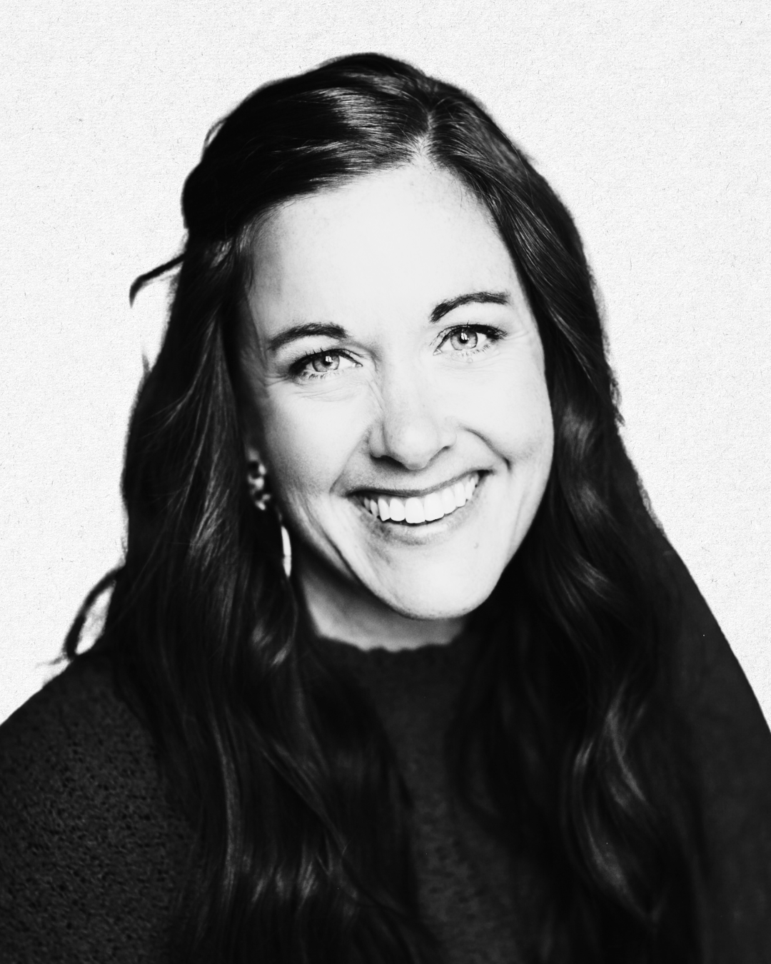 Black and white portrait of a woman with long dark hair, smiling and wearing earrings, against a plain background.