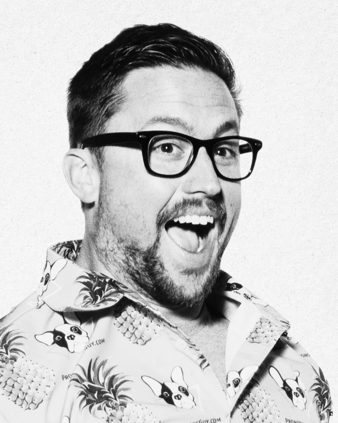 A black and white photo of a man with glasses, well-groomed hair and beard, mouth open in a cheerful expression, wearing a Hawaiian shirt with pineapple and dog face patterns.