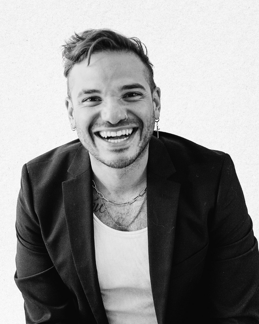 A smiling young man with earrings, wearing a black blazer over a white tank top, accessorized with necklaces, against a plain background.