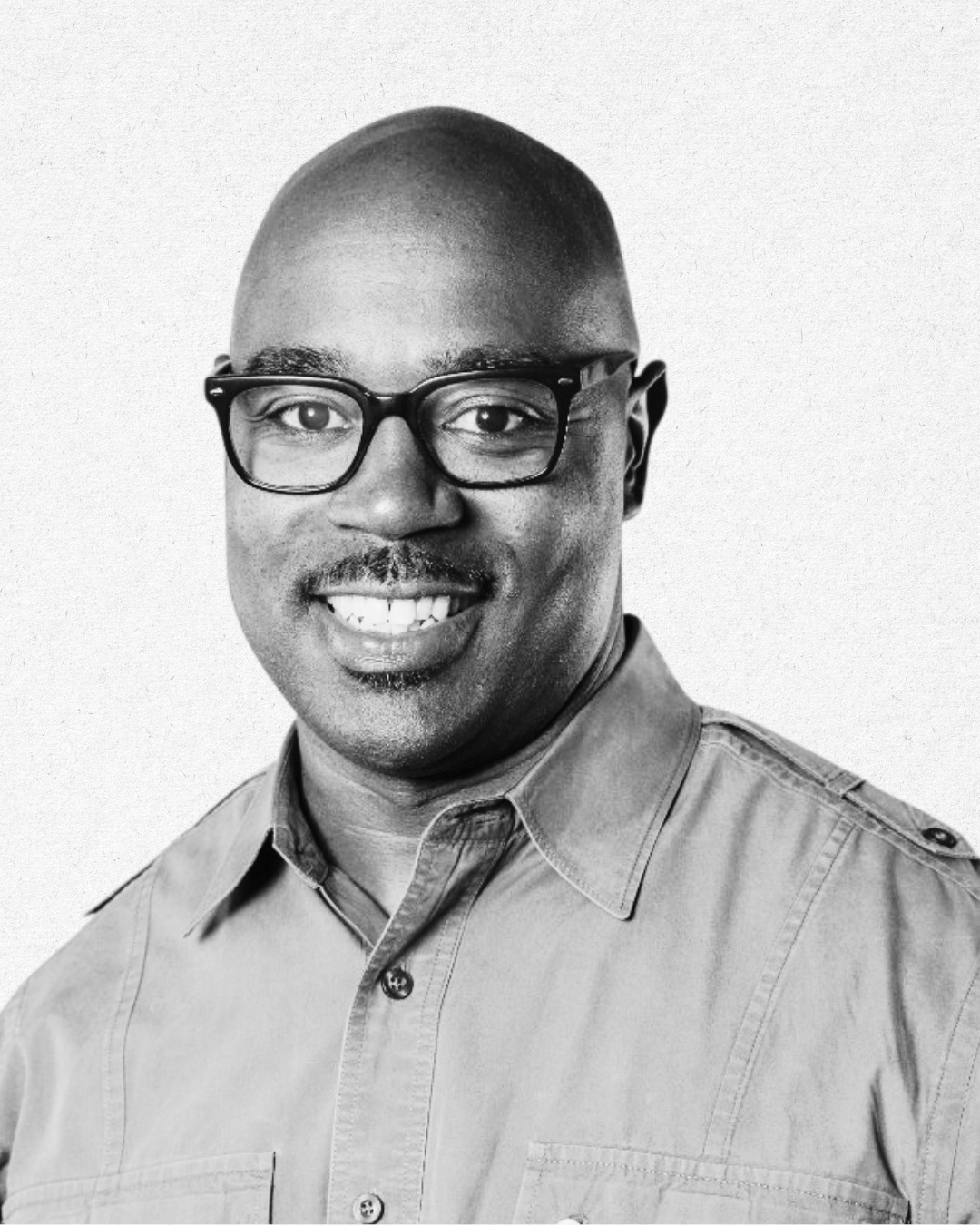 Black man wearing glasses and a collared shirt, smiling, in a black and white portrait.
