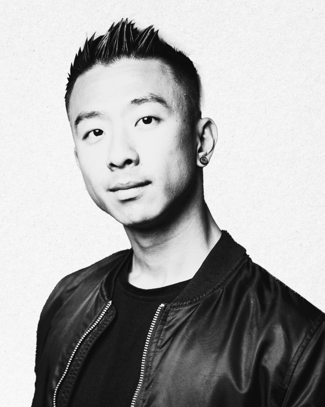 Black-and-white portrait of a young man with spiked hair, wearing a black sweatshirt and a black bomber jacket, against a plain light background.