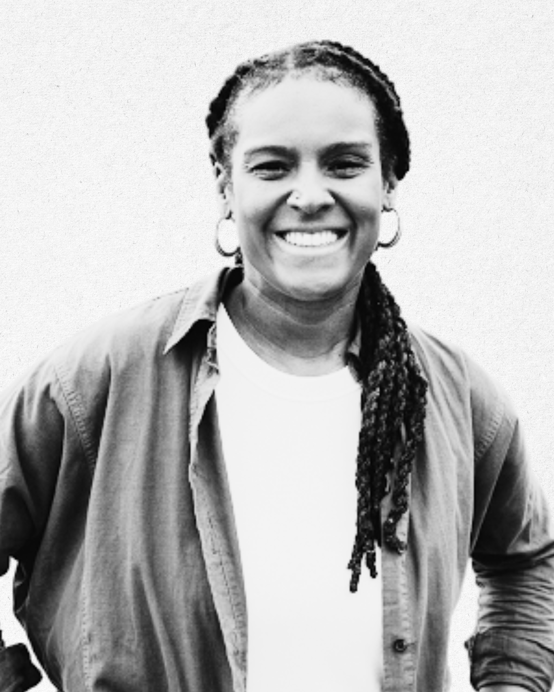 Black and white photo of a woman with dreadlocks, smiling, wearing hoop earrings, a light-colored shirt, and a dark jacket, standing against a plain background.