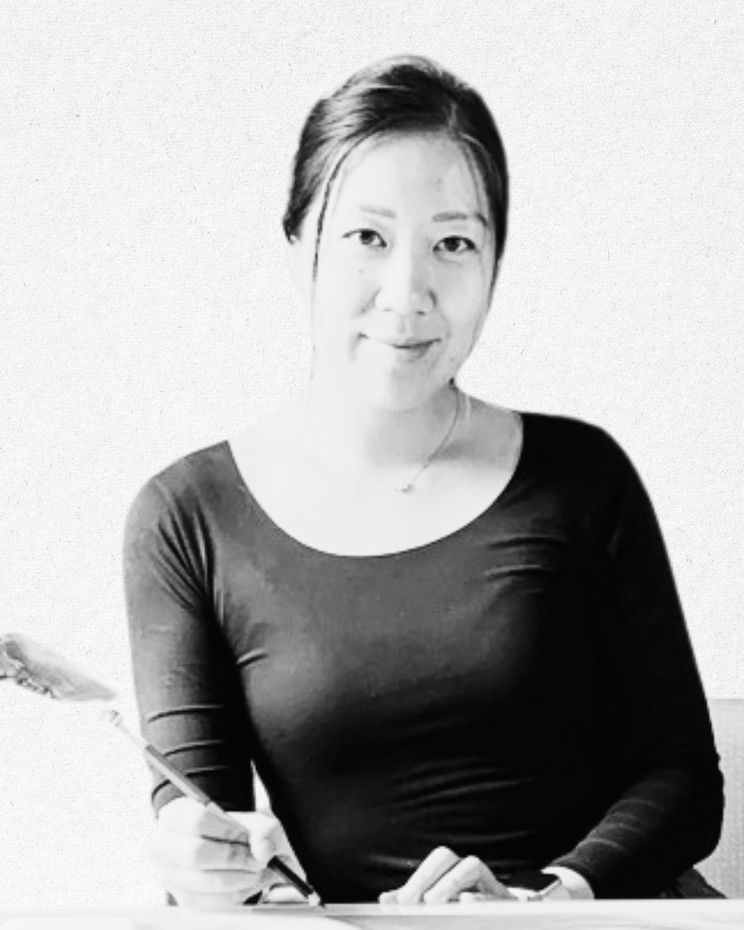 Black and white photo of a woman with short hair, wearing a black top, smiling slightly, sitting and holding a pen, with a plain background.