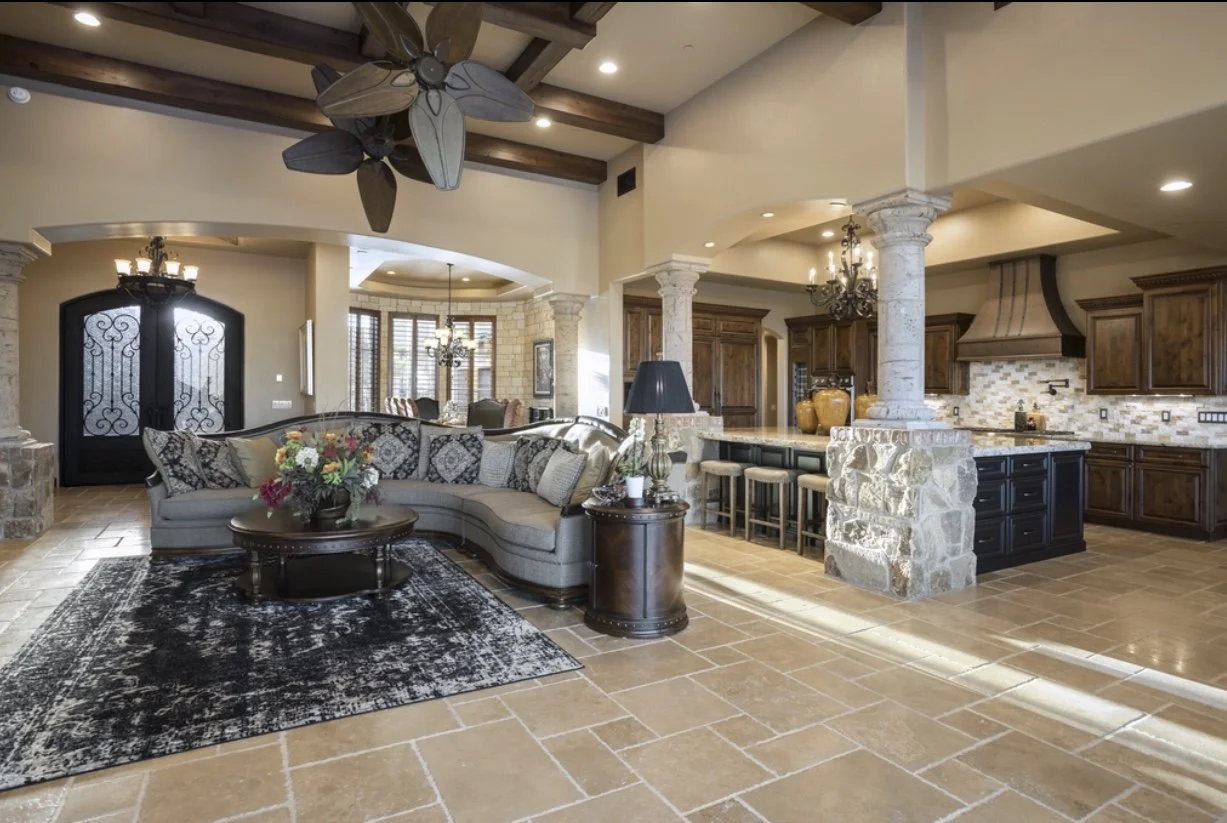 Luxurious open-concept great room with high ceilings crossed by dark wood beams and a large tropical-leaf ceiling fan. A curved gray sectional sofa with patterned pillows sits on a black distressed area rug around a round wood coffee table.