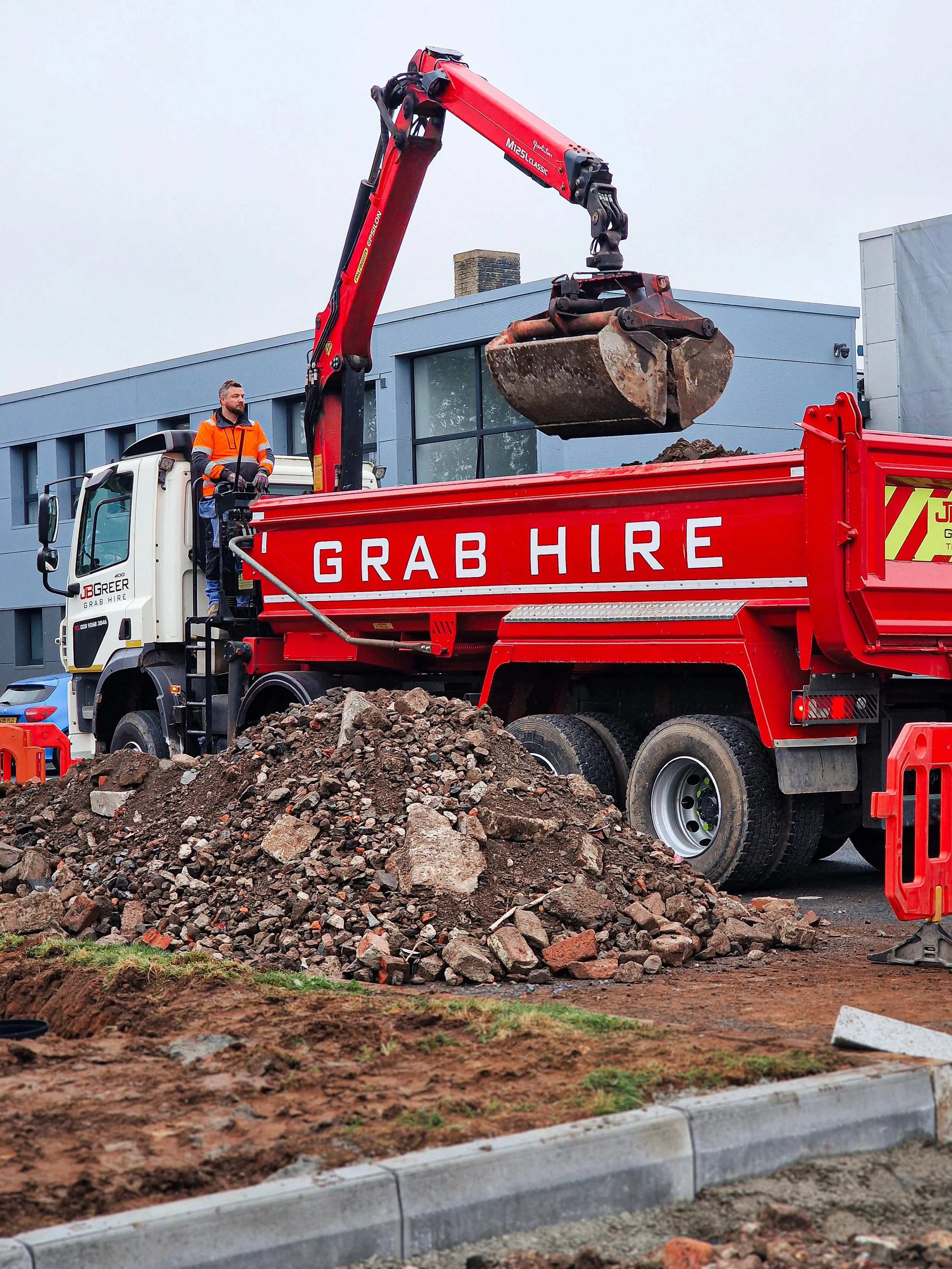 Construction worker in orange safety vest operating a red grab hire truck with a crane, lifting a bucket of dirt over a pile of rocks in front of a modern building.