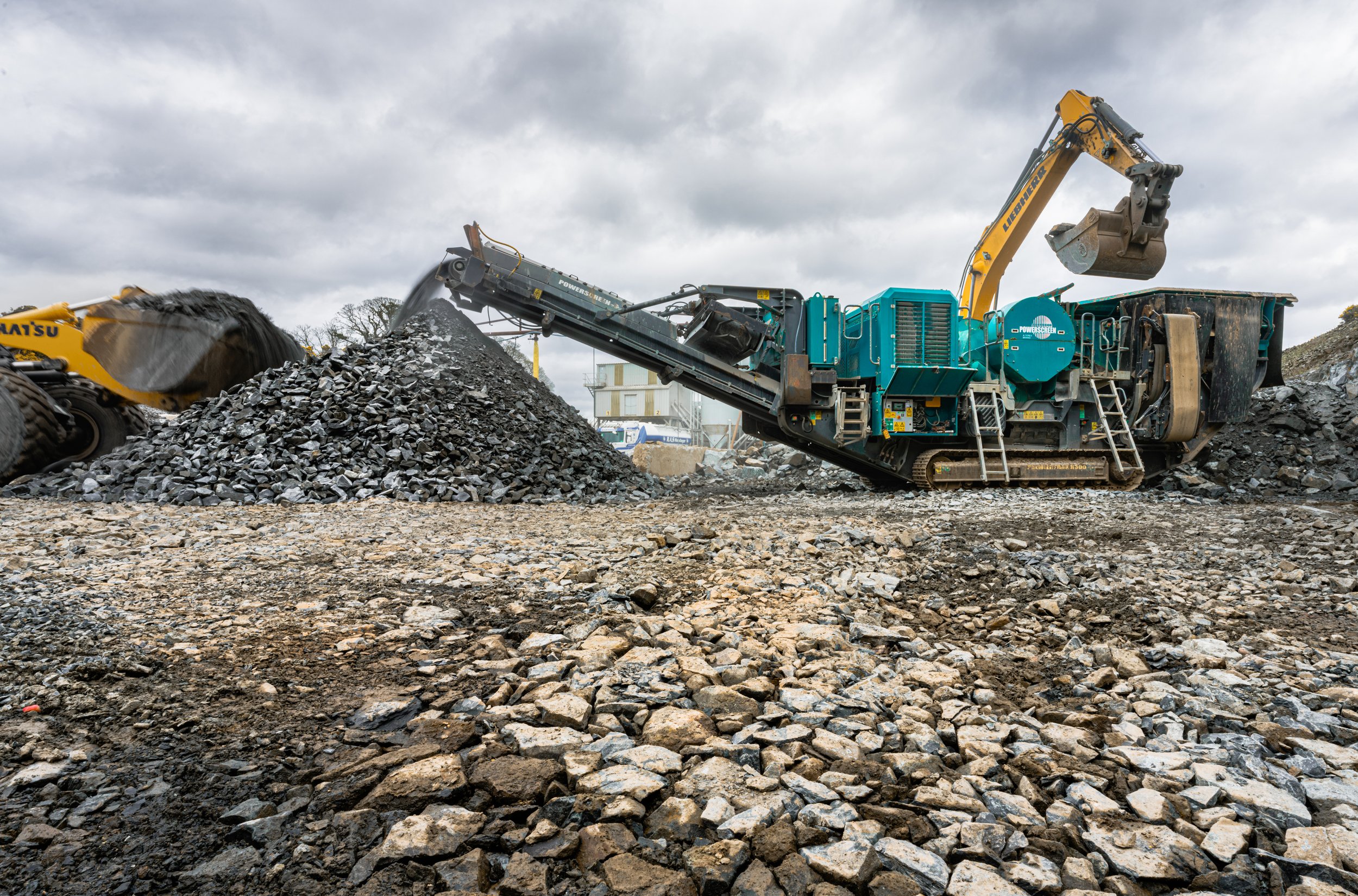 Construction site with a large crushermachine processing stone, with an excavator on the right and a pile of stone under a cloudy sky.