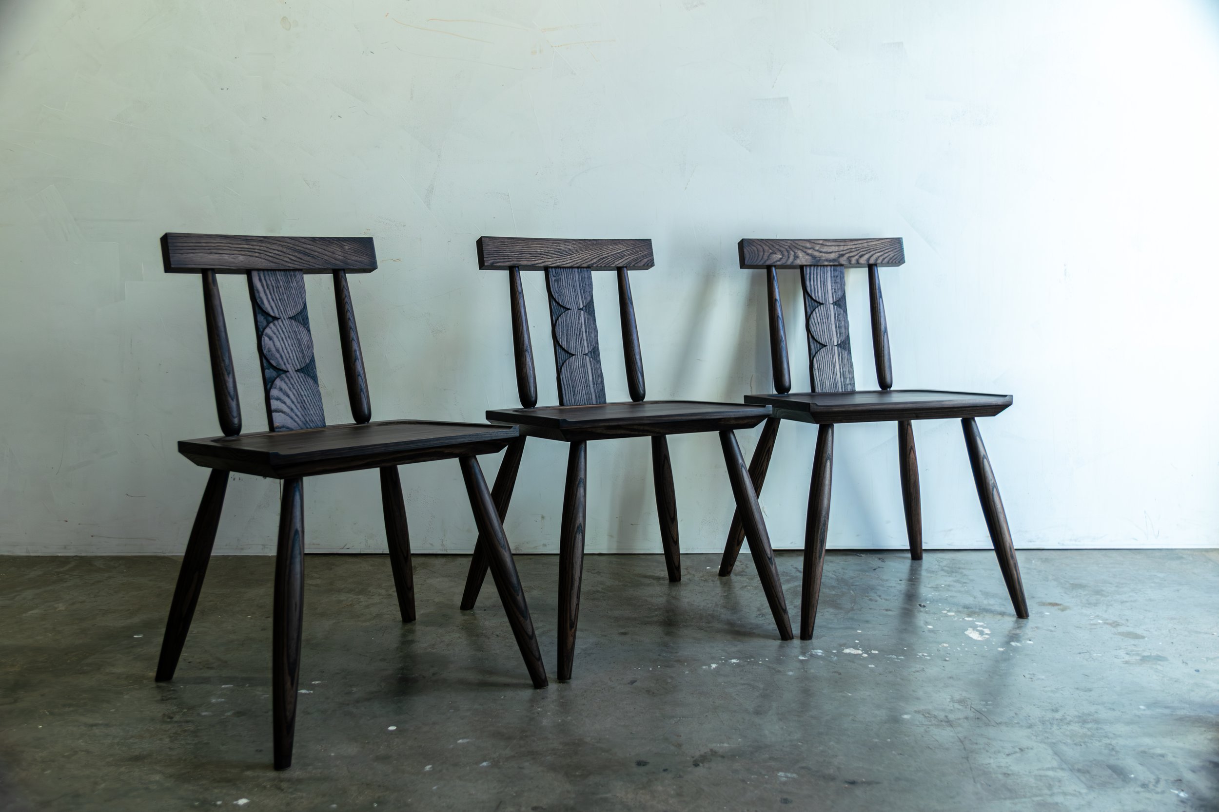 Three dark wooden chairs with backrests and angled legs are lined up against a plain white wall on a concrete floor.