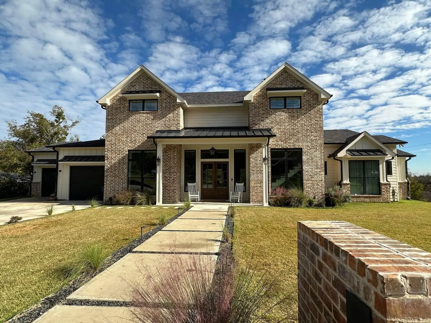Modern brick house with two stories, a black metal roof, and large windows. Features a front porch with rocking chairs, a garage, and a paved walkway leading through the lawn. Skies above are partly cloudy.