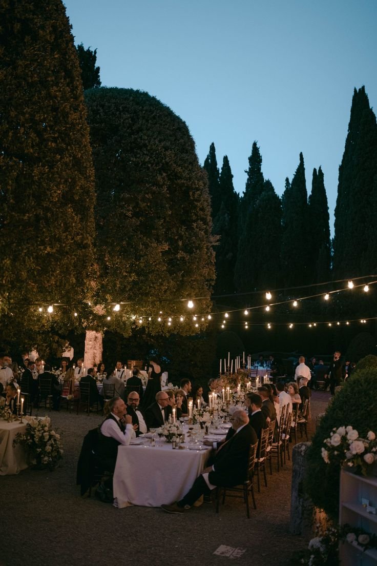 Outdoor evening wedding reception with guests seated at long tables, adorned with candles and string lights, set in a garden area with tall trees.