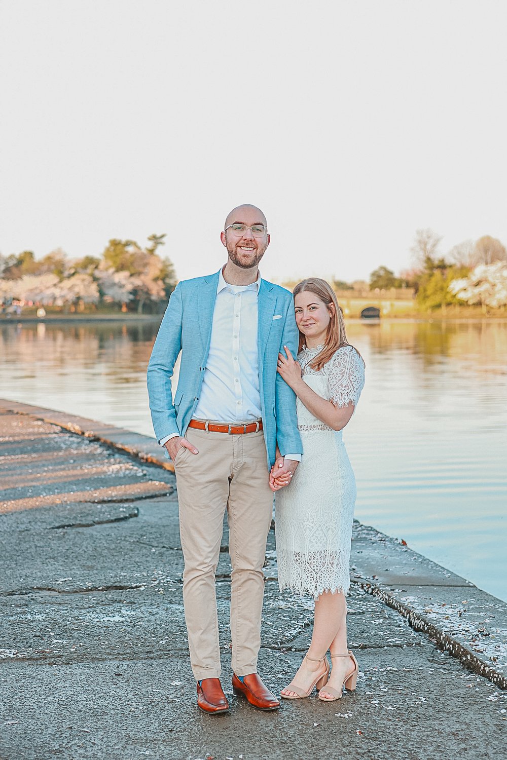 A couple posing by a waterfront with cherry blossom trees in the background. The man is wearing a light blue blazer and khaki pants, and the woman is in a white lace dress.