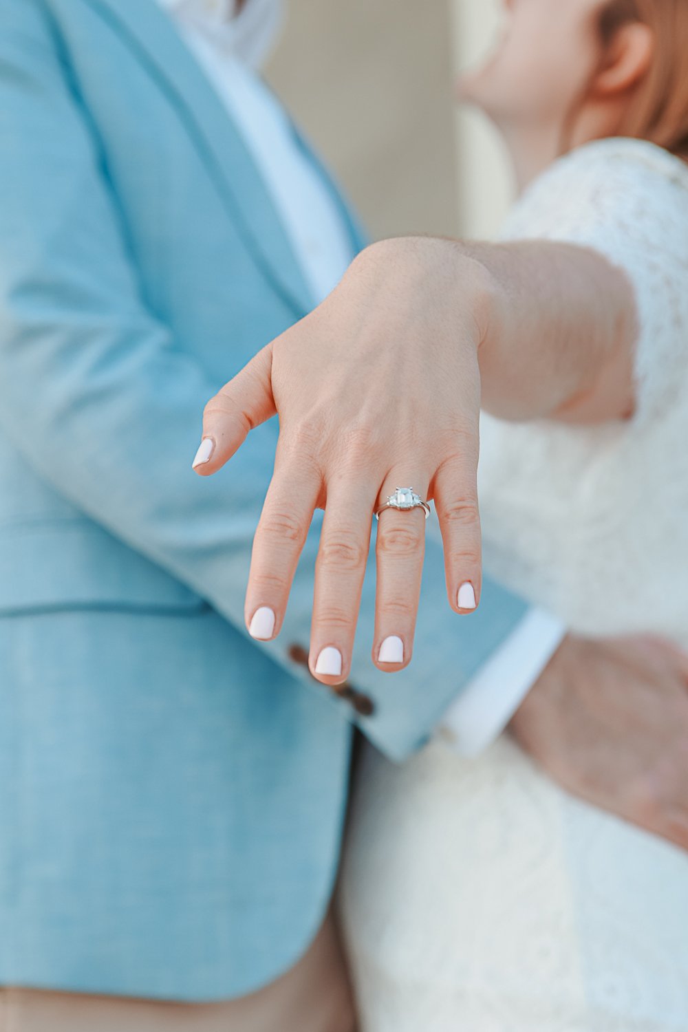 Close-up of a woman's hand with an engagement ring, focus on diamond, while a person in a blue suit stands in the background.