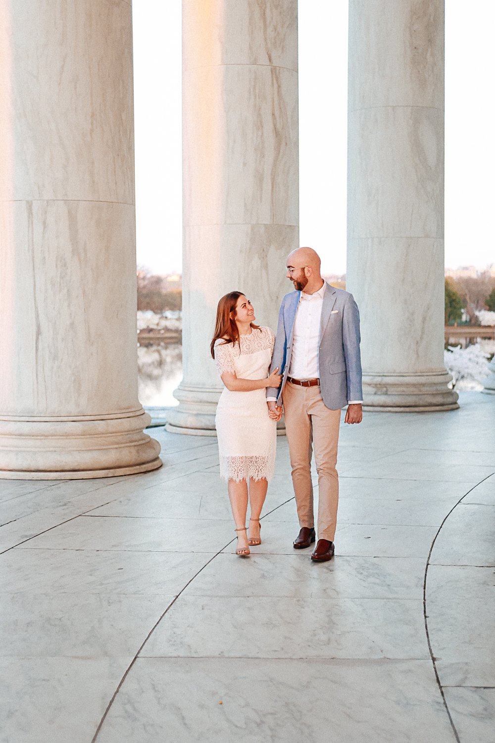 Couple walking between marble columns, dressed in formal attire, man in blue jacket and woman in lace dress.