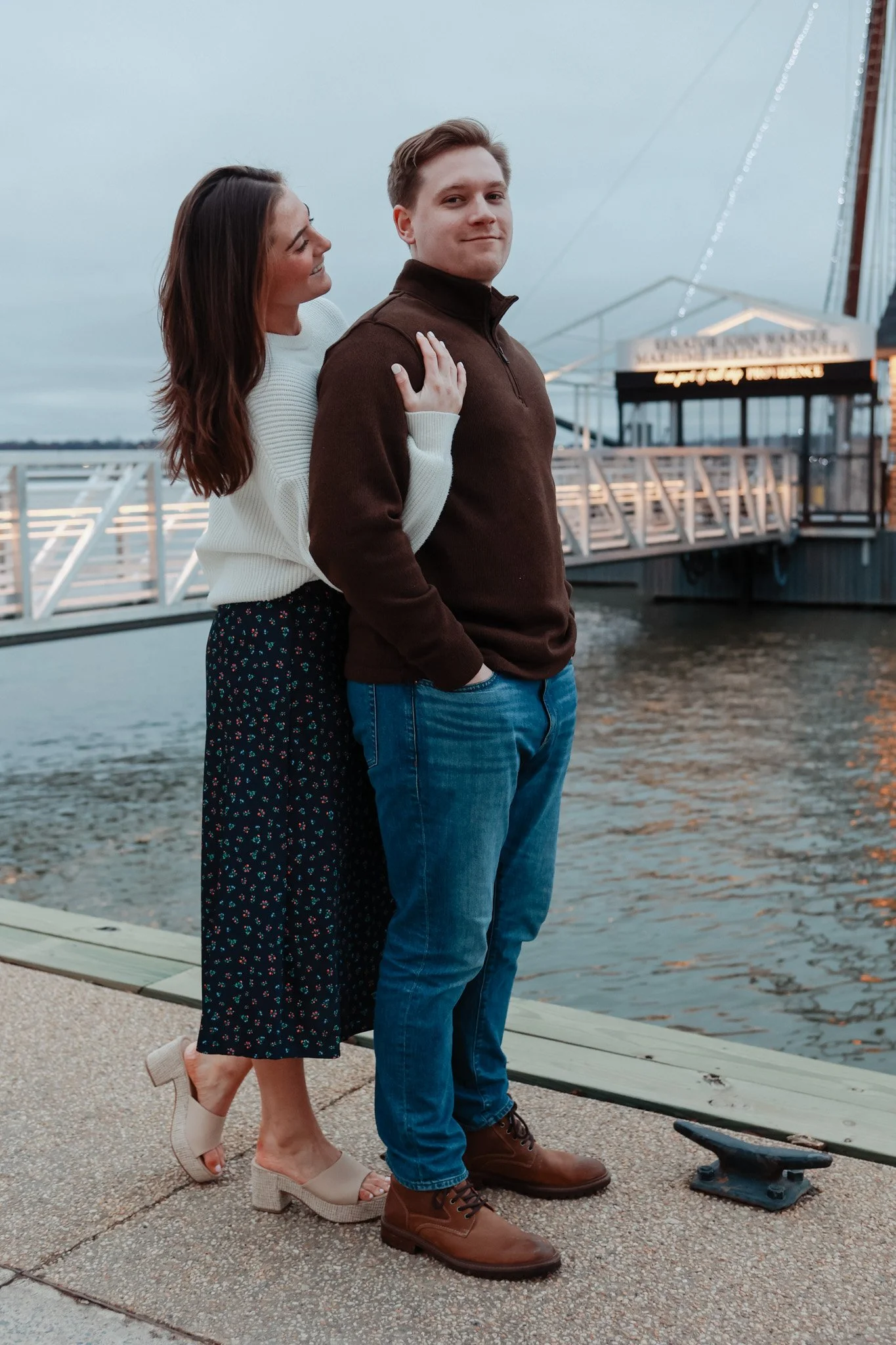 Couple standing on a dock near water, woman in white sweater and patterned skirt, man in brown sweater and jeans.