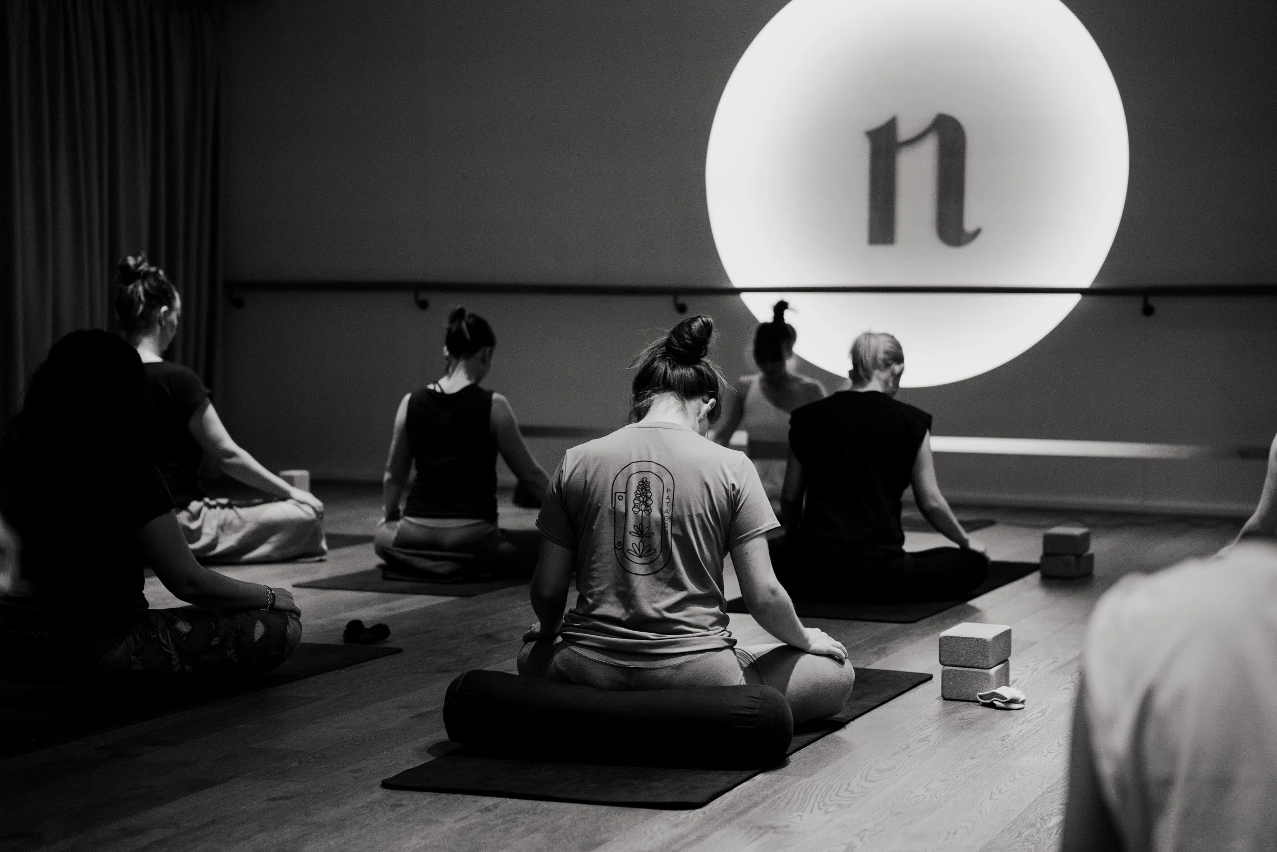 A group of women practicing yoga in a dimly lit studio, seated on mats with blocks and a towel beside them, facing a large illuminated screen displaying the letter 'n'.