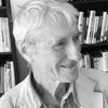 Black and white photo of an older man with white hair smiling, wearing a collared shirt and jacket, standing in front of bookshelves.