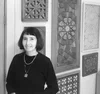 A woman standing in front of several geometric wood carvings on the wall, wearing a black top and necklace. The carvings exhibit intricate, symmetrical patterns.