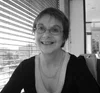 Smiling woman wearing glasses sitting indoors by window blinds in black and white photo.