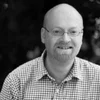Smiling man with glasses, bald head, and beard in a checkered shirt, black and white photo.